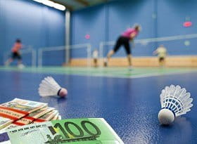 Im Vordergrund sind Geldscheine und zwei Federbälle und im Hintergrund finden Badmintonspiele statt.